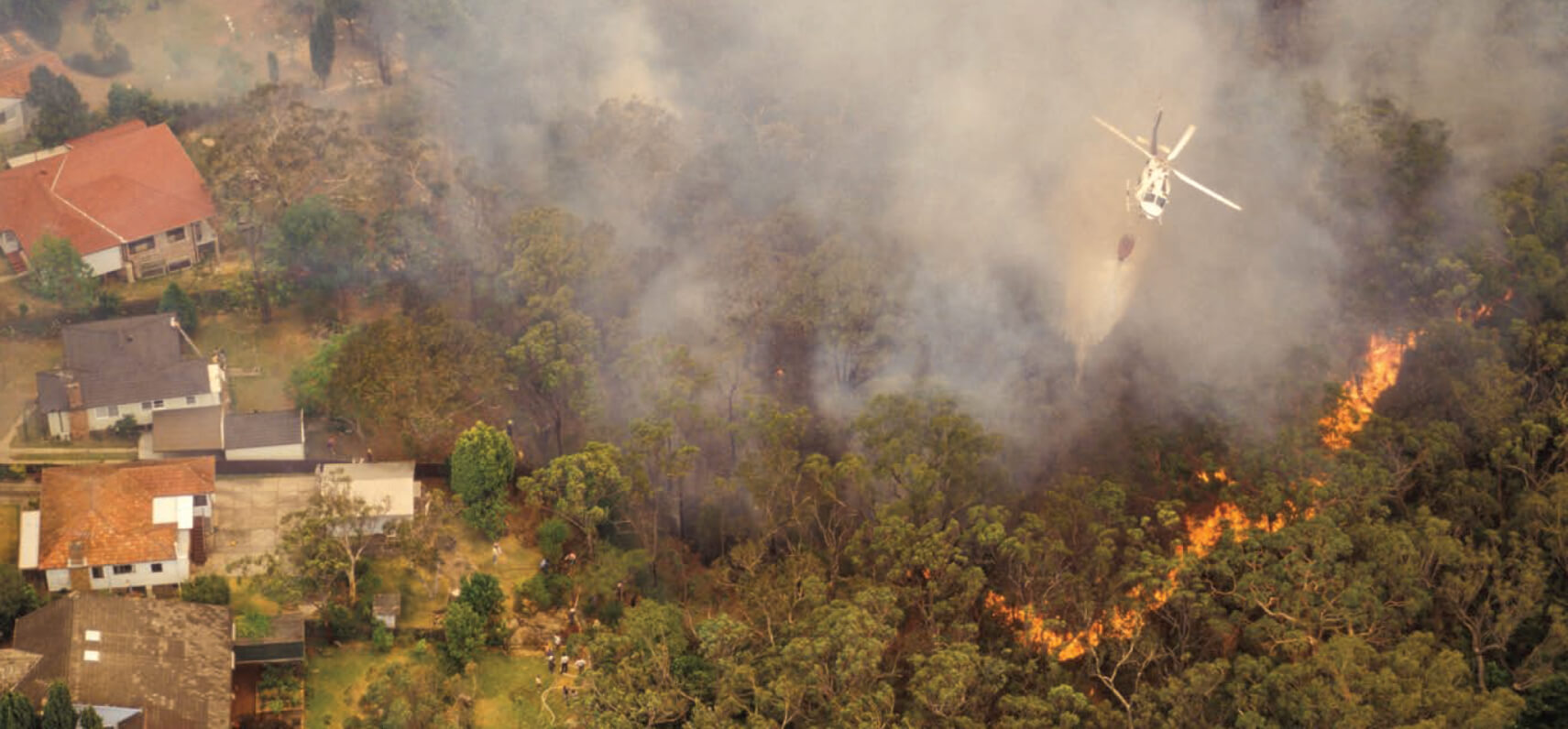 A high panorama shows a helicopter bombing water onto a burning line of fire in forest. To the left we see rows of house threatened by the flames.