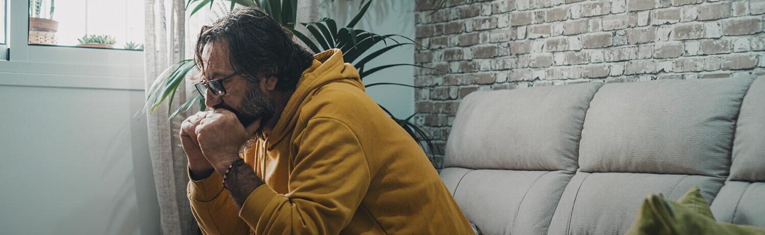A harried-looking man sits hunched forward on a couch, lost in his thoughts